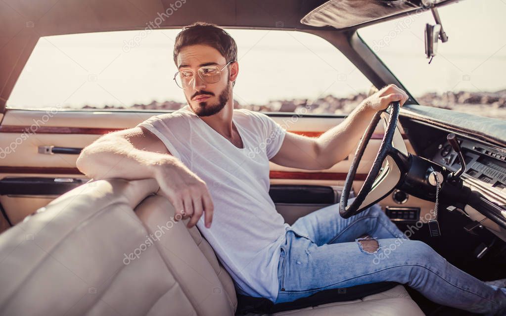 Handsome bearded man is sitting in his green retro car standing on the beach. Vintage classic car.
