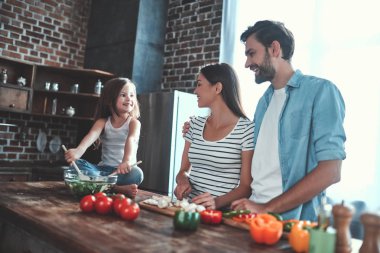 Anne, baba ve kızı mutfakta yemek pişiriyor. Mutlu aile kavramı. Yakışıklı adam, çekici genç kadın ve tatlı küçük kızları birlikte salata yapıyorlar. Sağlıklı yaşam tarzı.