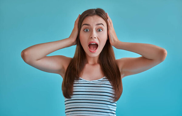 Attractive young woman isolated on blue background. Portrait of girl screaming with ears closed by hands.
