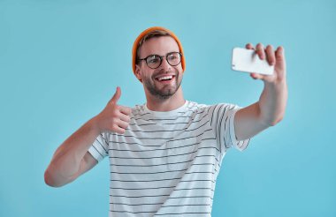 Yakışıklı adam izole. Gözlük şık genç adam mavi arka plan üzerinde poz ve selfie üzerinde akıllı telefon görüşmesi.