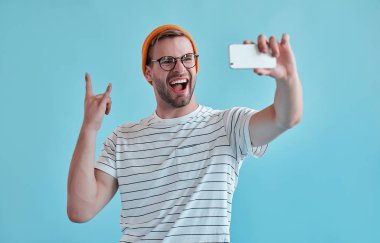 Yakışıklı adam izole. Gözlük şık genç adam mavi arka plan üzerinde poz ve selfie üzerinde akıllı telefon görüşmesi.