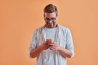 Yakışıklı adam izole. Gözlük şık genç adam akıllı telefon turuncu arka plan ile poz. Kulaklık kullanarak müzik