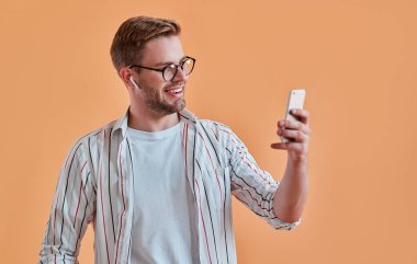 Yakışıklı adam izole. Gözlük şık genç adam akıllı telefon turuncu arka plan ile poz. Kablosuz kulaklık kullanarak müzik