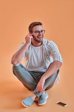 Yakışıklı adam izole. Gözlük şık genç adam akıllı telefon turuncu arka plan ile poz. Kablosuz kulaklık kullanarak müzik