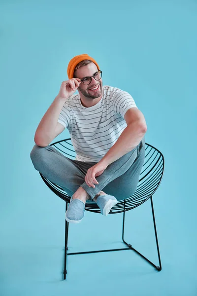 Handsome man isolated. Stylish young man in eyeglasses is  posing while sitting on chair on blue background.