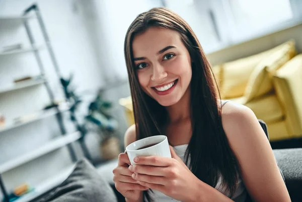 Good morning! Attractive young woman is having rest at home. Girl sitting on a sofa with cup of coffee and smiling.