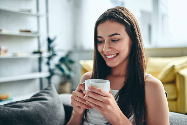 Доброе утро! Привлекательная молодая женщина отдыхает дома. Девушка сидит на диване с чашкой кофе и улыбается
.