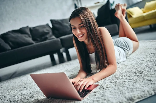 Good morning! Attractive young woman is having rest at home. Girl lying on a carpet with laptop.