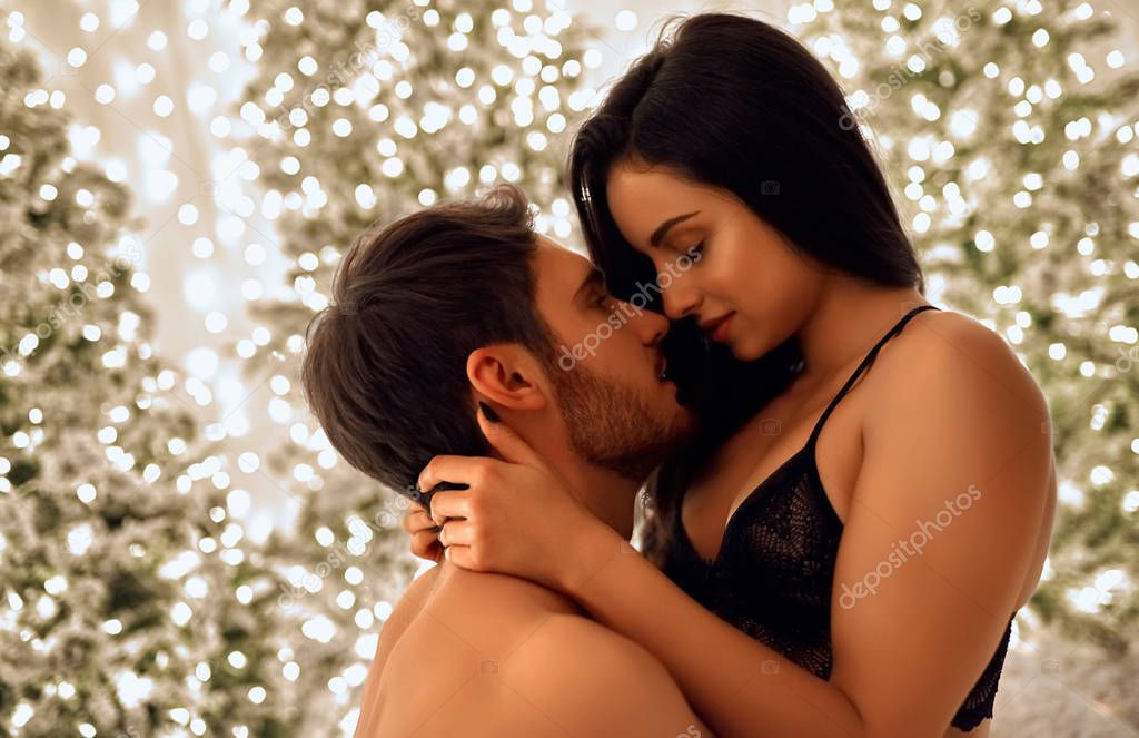 Merry Christmas and Happy New Year! Sexy passionate couple celebrating Christmas together. Attractive woman in lingerie and handsome bearded man having sex on background of decorated New Year trees.