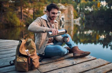 Erkek turist yeni yerler keşfetmek. Sakallı yakışıklı niteliğine. Termos ile oturma ve göl kenarında çay içme.