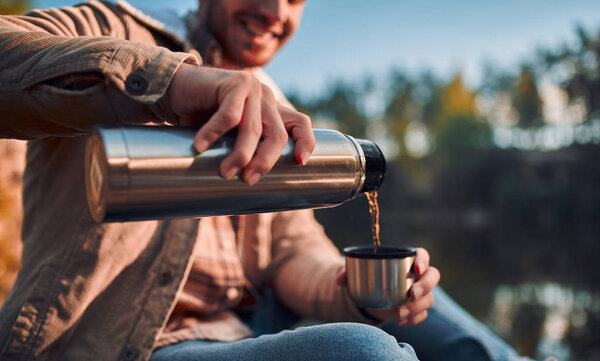 Male tourist exploring new places. Handsome bearded man on nature. Sitting with thermos and drinking tea near lake.