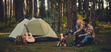 Young couple of tourists are exploring new places. Attractive woman and handsome man are spending time together on nature. Sitting near bonfire and touristic tent in forest and making marshmallow. clipart