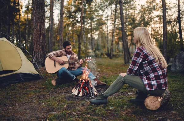 Young couple of tourists are exploring new places. Attractive woman and handsome bearded man are spending time together on nature. Sitting near bonfire and touristic tent in forest and playing guitar.