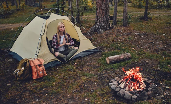 Female tourist exploring new places. Attractive young woman spending time on nature. Sitting and meditating in touristic tent in forest near bonfire.