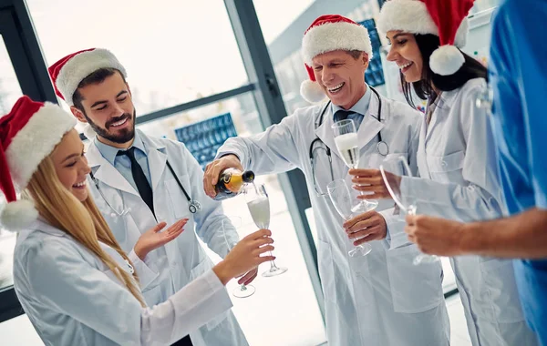 Merry Christmas and Happy New Year! Group of doctors celebrating winter holidays at work. Medical personnel in uniform and Santa Claus hats drinking champagne together.