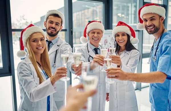 Merry Christmas and Happy New Year! Group of doctors celebrating winter holidays at work. Medical personnel in uniform and Santa Claus hats drinking champagne together.