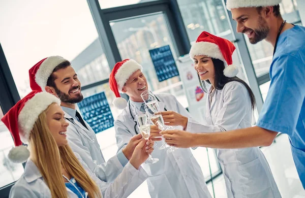 Merry Christmas and Happy New Year! Group of doctors celebrating winter holidays at work. Medical personnel in uniform and Santa Claus hats drinking champagne together.