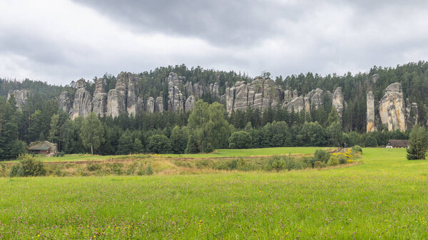 Adrspach-Teplice Rocks, sandstone formations in Hradec Kralove Region in Czech Republic, Europe. A dramatic landscape of unique rock formations amidst a lush green meadow, under a cloudy sky above.