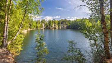 Çek Cumhuriyeti 'nin Avrupa' daki Hradec Kralove Bölgesi 'ndeki Adrspach-Teplice Kayalıkları bölgesinde bulunan Piskovna Gölü. Parlak mavi gökyüzünün altında yemyeşil orman ve kumtaşı oluşumlarıyla çevrili manzaralı bir göl..