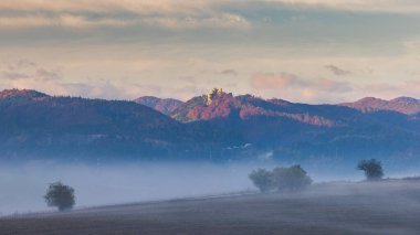 Slovakya, Avrupa 'daki Lietava şatosu. Sonbahar manzarası, tarlaların üzerinde sis ve gündoğumunda tepenin üzerindeki uzak şato. Doğanın manzaralı güzelliği.