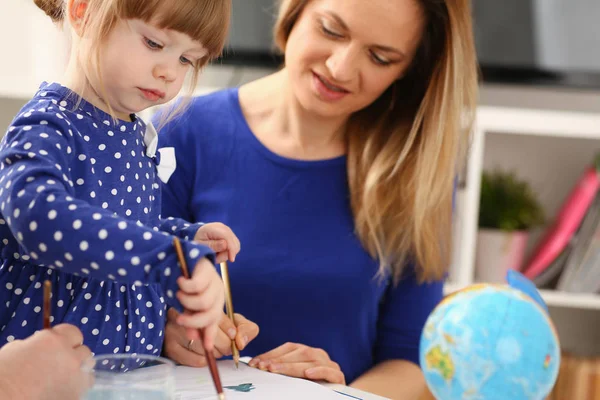 Blond smiling little girl hold in arm brush drawing something together with mom. Beautiful female young artist, minor baby-sitter, art joy, juvenile development, parent lifestyle, youth, painter