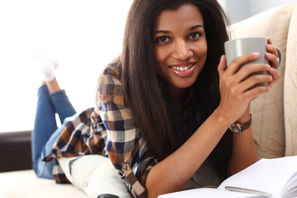 Smiling black woman lie down at sofa with cup of hot beverage Royalty Free Stock Images