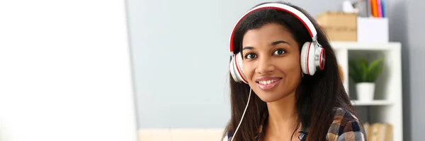 Preto sorrindo mulher sentada no local de trabalho usando fones de ouvido — Fotografia de Stock