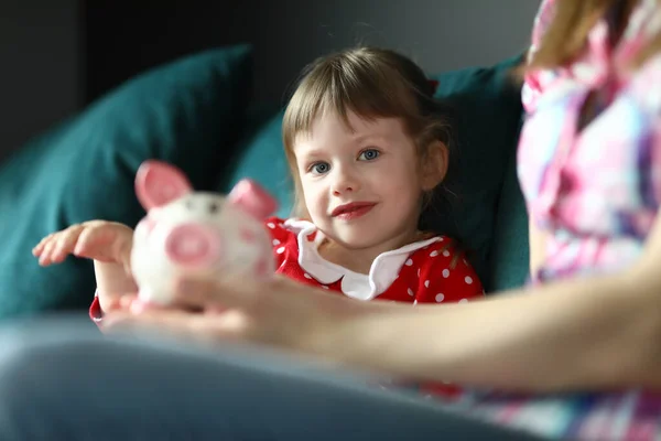 Mom and daughter sit home on couch with piggy bank