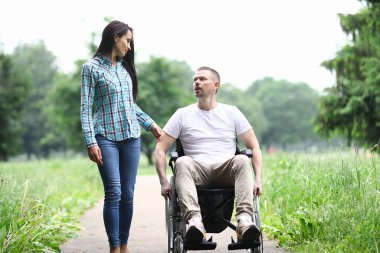 Tekerlekli sandalyedeki kadın ve adam parkta yürüyüp konuşuyorlar.