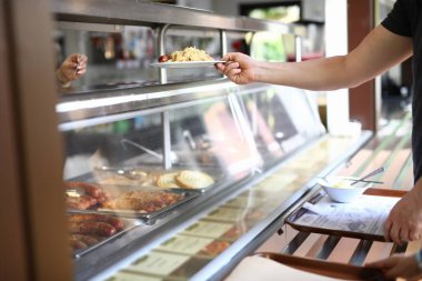 Adam yemek odasında hazır yemek tezgahının arkasında duruyor ve elinde öğle yemeğiyle tabağı tutuyor..