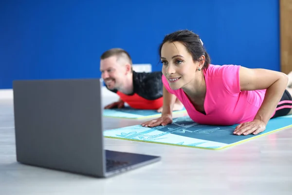 Woman and man do push-up on gymnastic mat and watch video in loptop.