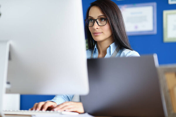 Woman in glasses and blue shirt sit at workplace and look at computer screen.