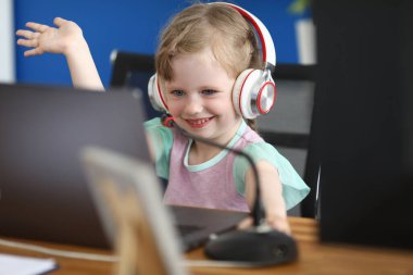 Küçük kız mikrofonlu kulaklıkla bilgisayarın başına oturur, gülümser ve elini sallar.