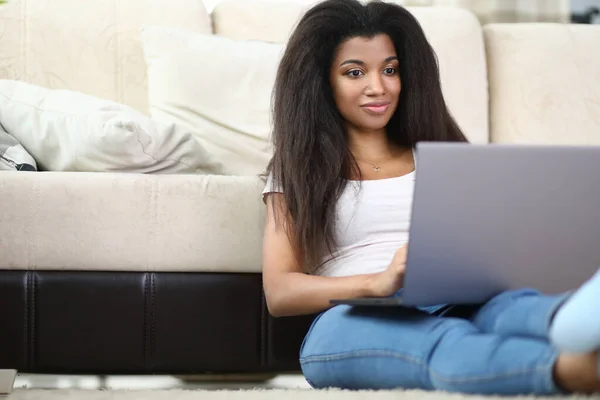 Femme africaine est assis à la maison à côté du canapé et tient ordinateur portable. — Photo