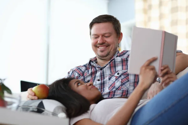 Mannen sitter på soffan leende på knä kvinna ligger och håller bok i sina händer. — Stockfoto