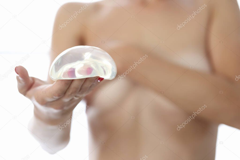 Nude woman holds breast implant in hand