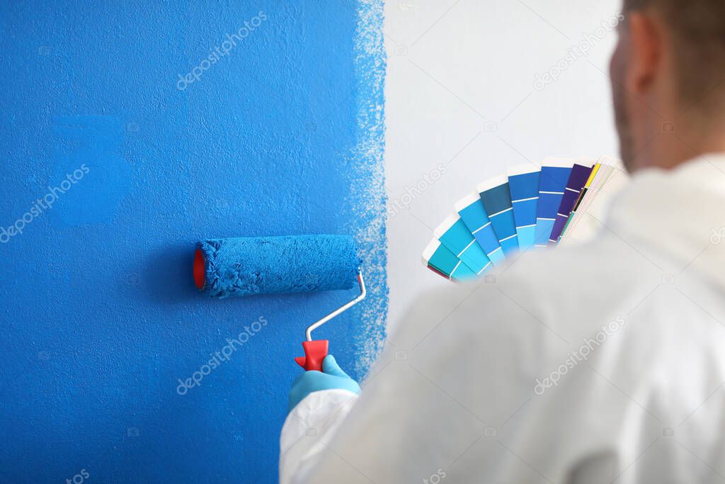 Craftsman holds roller and a color palette and paints white wall blue