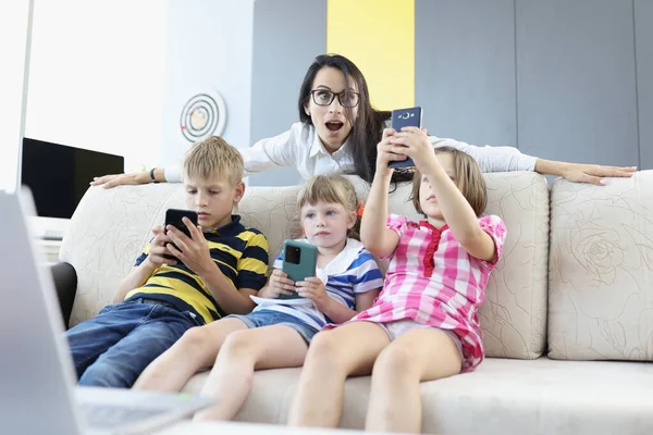 Tres niños están sentados en el sofá con teléfonos inteligentes en sus manos, jugando juegos en línea con la mujer de pie detrás del sofá y mirando asustados en la pantalla del teléfono. — Foto de Stock