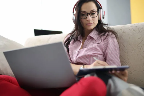 Mulher em fones de ouvido com área de transferência em suas mãos senta-se no sofá com laptop — Fotografia de Stock