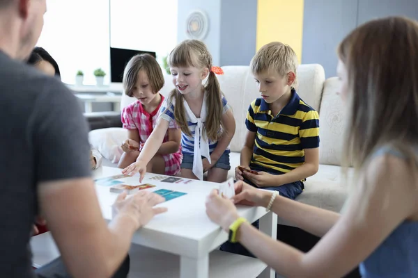 Adults and children are sitting around table on which playing cards are located girl reaches for card with her hand. — Stock Photo, Image