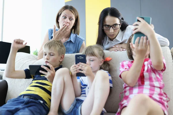 Tre barn sitter på soffan med smartphones i händerna, spelar online-spel från baksidan av soffan, två kvinnor står och ser rädda ut på telefonskärmen. — Stockfoto