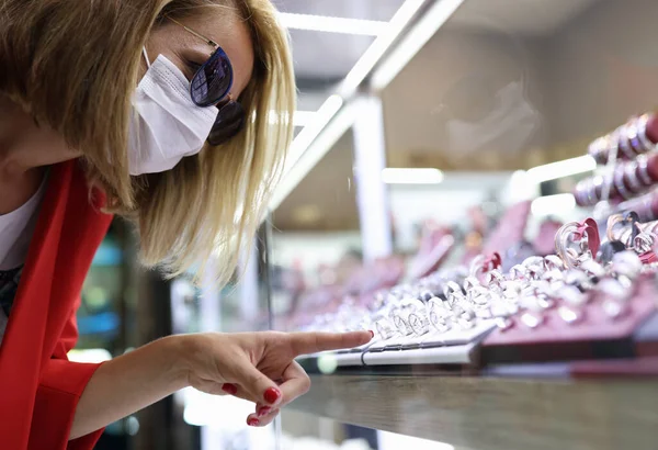 Jonge vrouw met medisch masker, zonnebril, rode jas wijst haar vinger naar sieraden display — Stockfoto