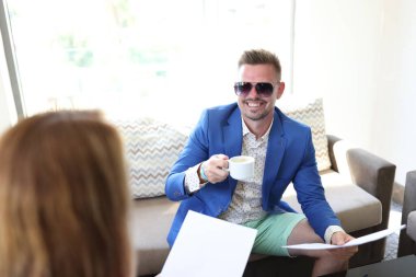 Kahvesinde bir fincan kahve ve belgeyle mavi takım elbiseli, gülümseyen genç bir adam kafede bir kadınla buluşuyor.