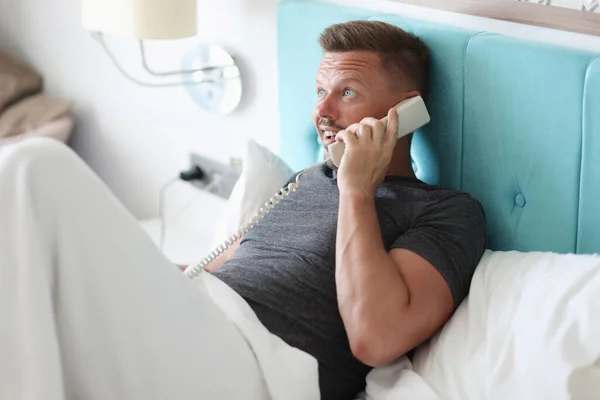 Чоловік лежить в ліжку в готельному номері і розмовляє по телефону . — стокове фото