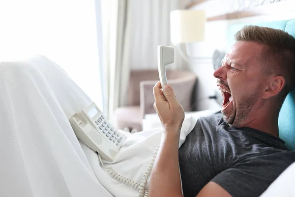 Angry man lies in bed and shout into white phone receiver.