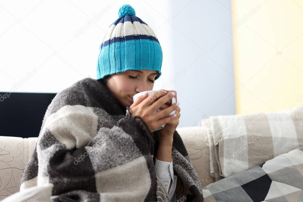 Sick woman in hat under blanket holds cup of medicine