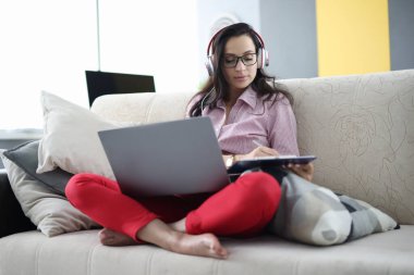 Kulaklıklı bir kadın dizüstü bilgisayarla koltukta oturuyor ve elinde bir pano tutuyor.