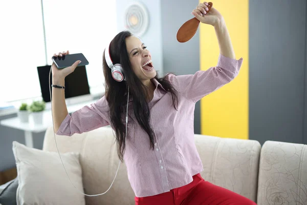 Kobieta w słuchawkach na kanapie trzyma smartfona w rękach i udaje, że śpiewa — Zdjęcie stockowe