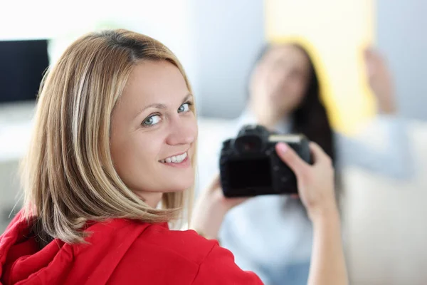 Młoda kobieta fotograf trzyma aparat fotograficzny w dłoniach z brunetką w tle w domu — Zdjęcie stockowe