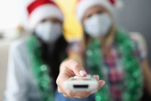 Les femmes dans les chapeaux de Noël et les masques de visage tiennent une télécommande TV — Photo